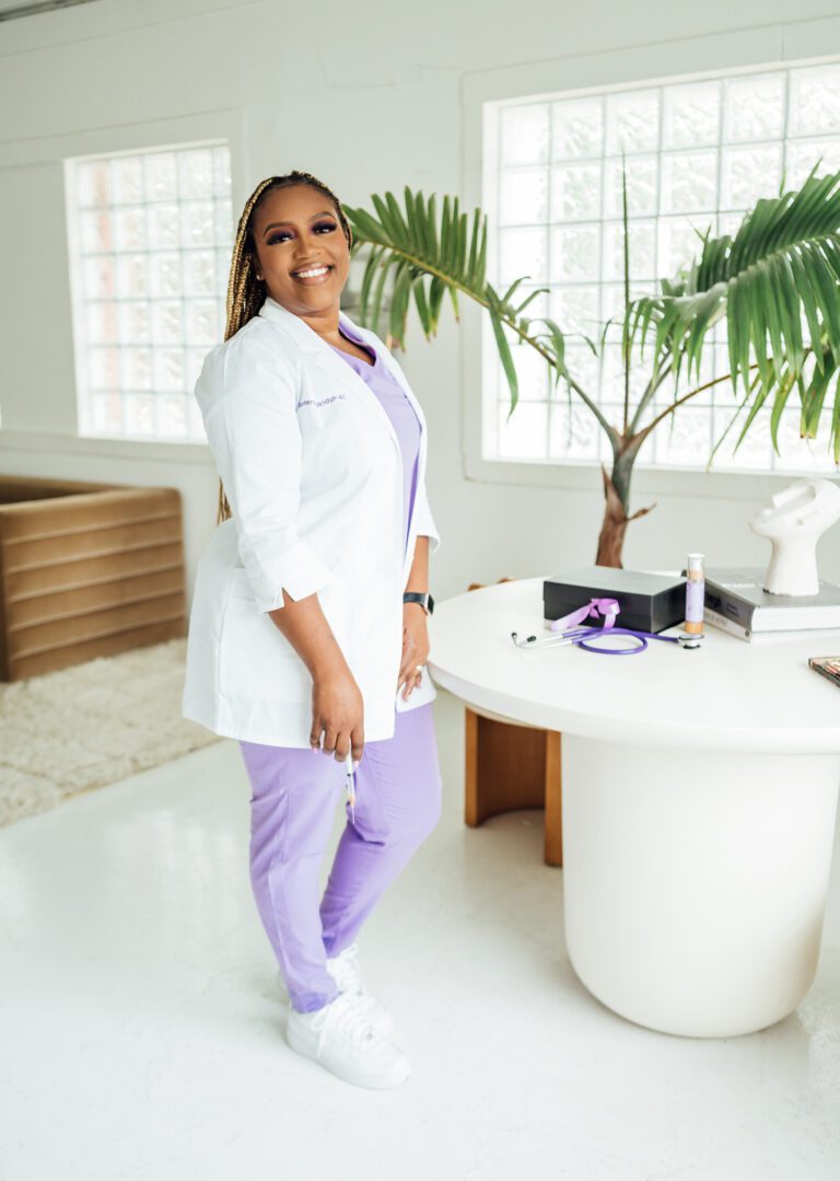 Smiling female medical professional in a white coat and purple scrubs standing in a modern office setting.