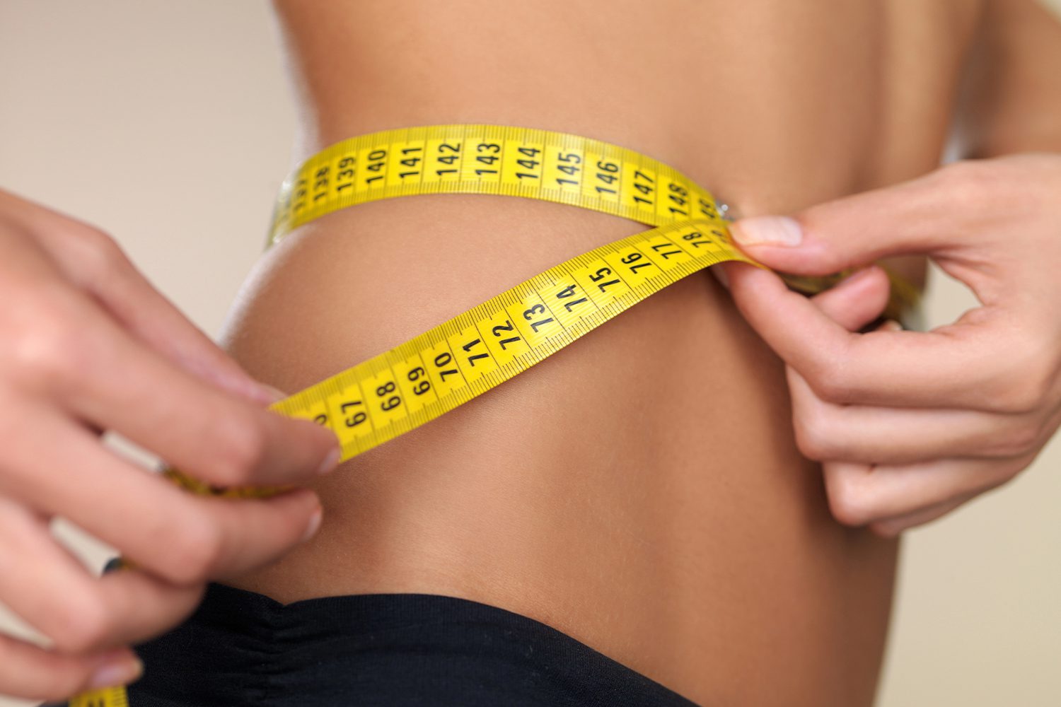 Close-up of a measuring tape around a woman's waist as she tracks her weight loss progress.