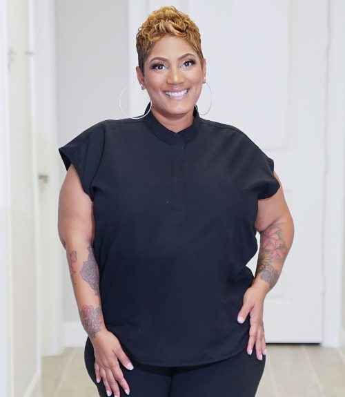 Smiling woman in black outfit with tattoos standing confidently in a modern indoor setting.